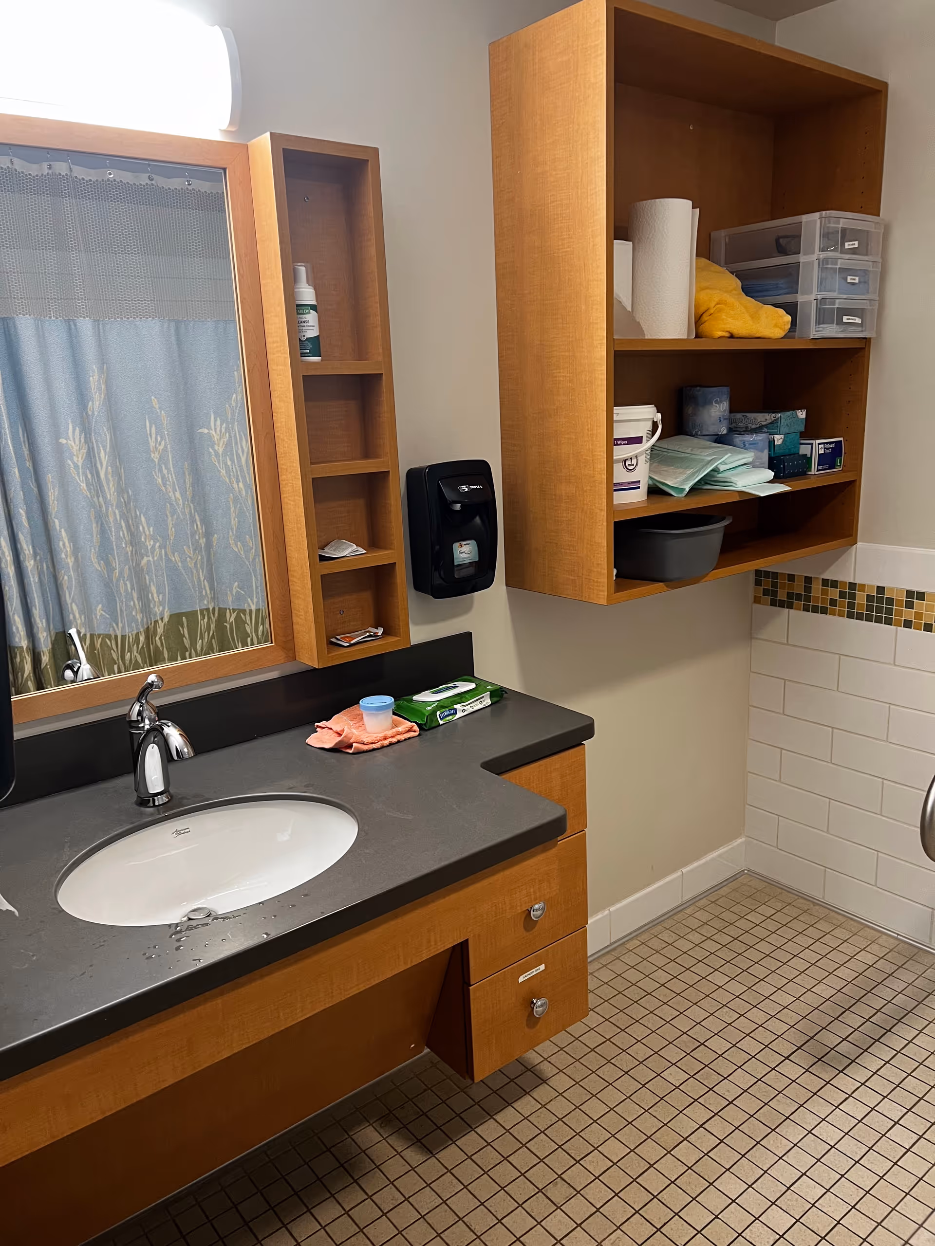 Bathroom with sink, counter, shelves, mirror, and tiled floor in a nursing facility.
