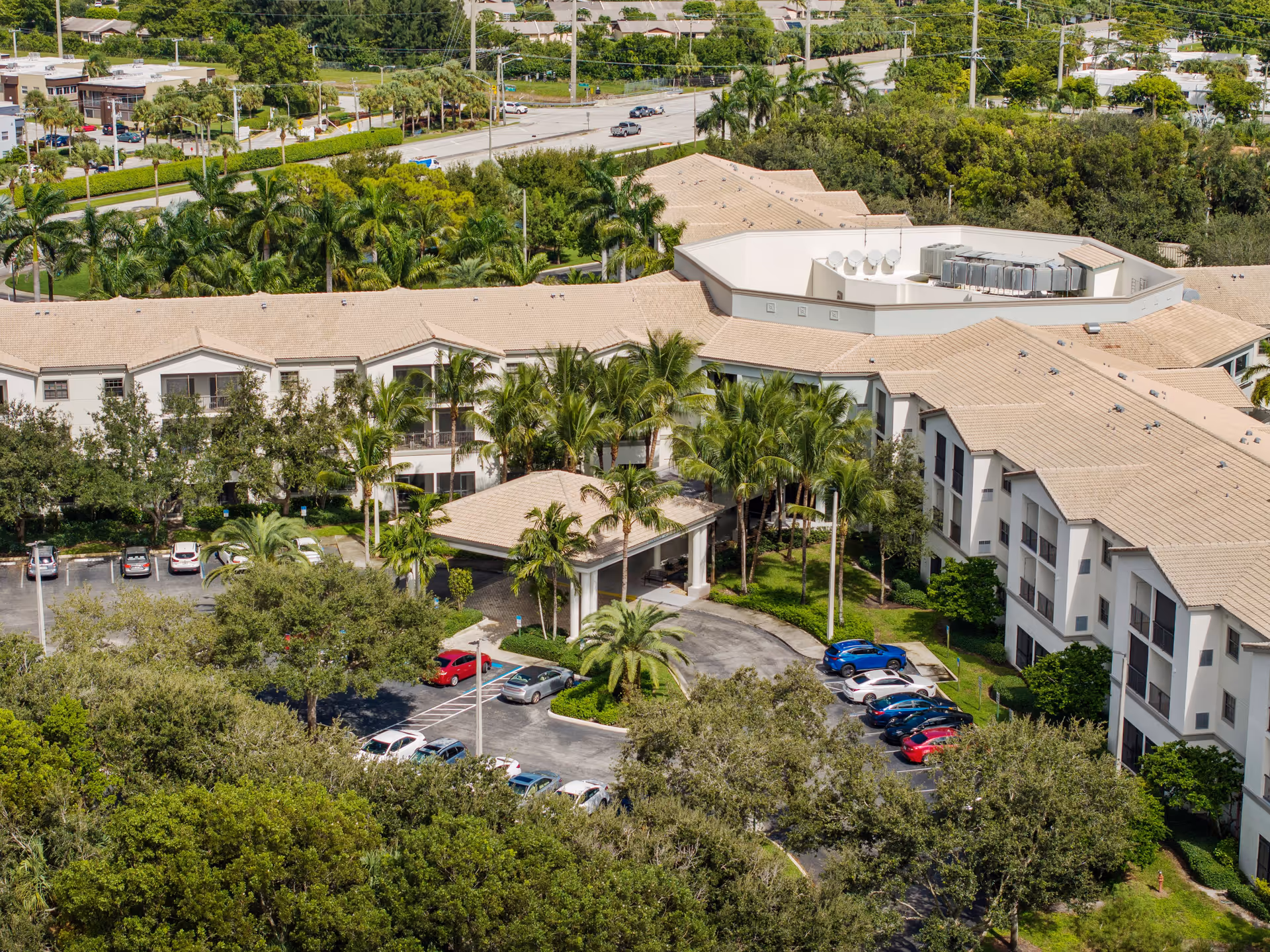 Aerial view of Verena at Delray senior living facility showing a large, multi-wing building with beige roofs surrounded by palm trees and greenery. There is a covered entrance with a driveway and several parked cars in the parking lot. Roads and additional greenery are visible in the background.