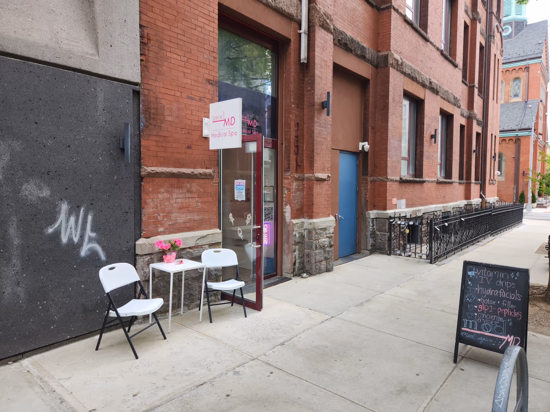 Exterior brick building with sidewalk seating, small table, two white chairs, and chalkboard sign outside.