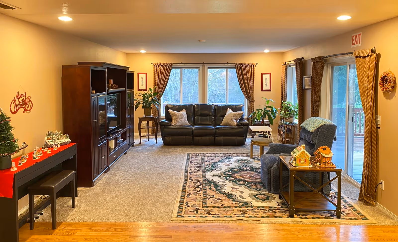 A cozy living room with a black leather sofa against a wall with three large windows covered by brown curtains. There is a patterned area rug on the carpeted floor, a blue armchair with a side table holding decorative gingerbread houses, and several potted plants near the windows. On the left side, there is a dark wood entertainment center and a small piano with Christmas decorations on top. The walls are painted beige, and there is an exit sign above a sliding glass door on the right.