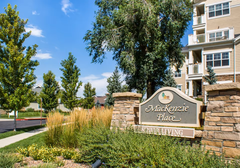 Stone entry sign reading "MacKenzie Place" surrounded by landscaping in front of a multi-story senior living building.