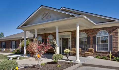 Front exterior view of Churchill Terrace Senior Living building with brick walls, white columns supporting a covered entrance, arched windows, outdoor seating, and landscaped garden with small trees and shrubs under a clear blue sky.