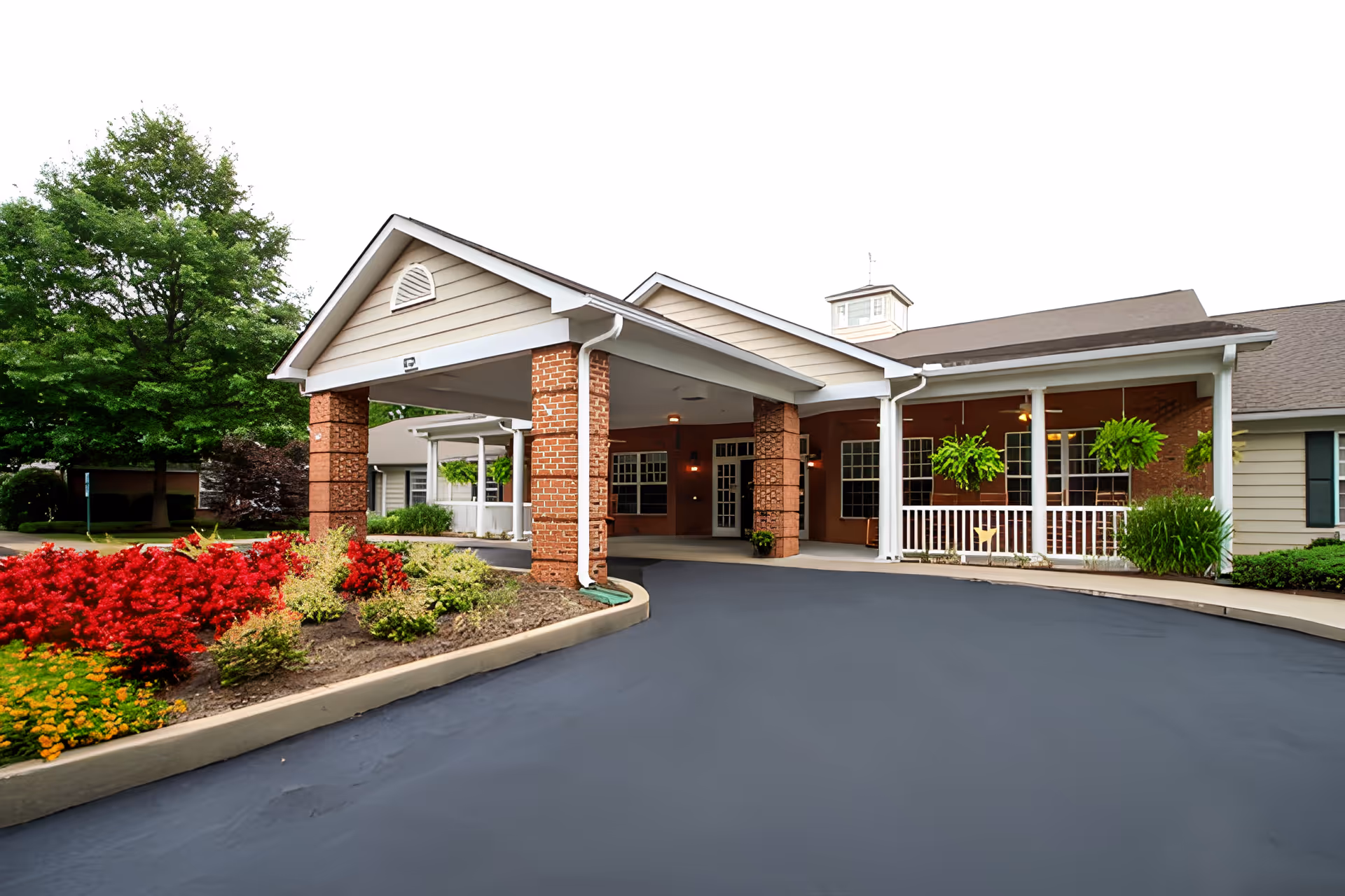 Front entrance of a senior living facility with a covered drop-off area supported by brick columns. The building has beige siding and a gray roof. There are hanging plants and a landscaped garden with red and yellow flowers near the driveway.