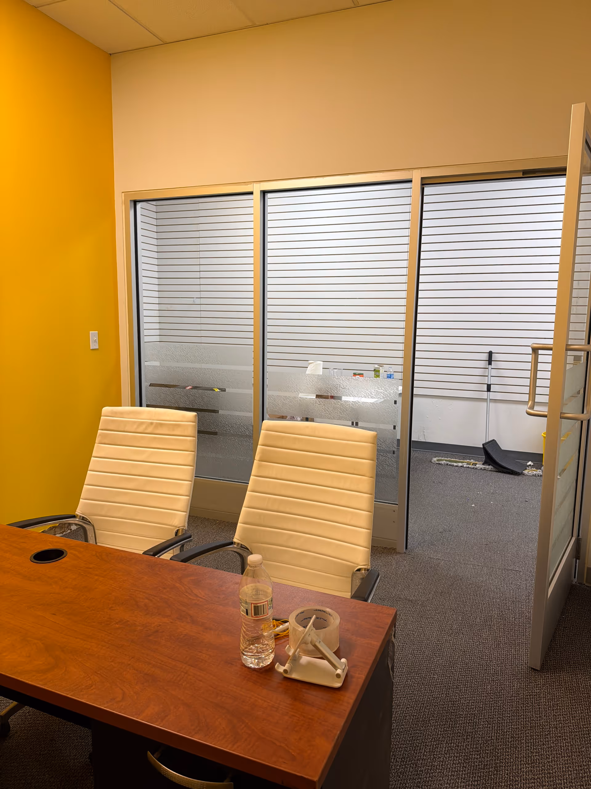 Yellow-walled conference room with glass partition, wooden table, and beige chairs.