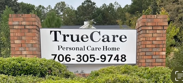 A large white sign with black text reading 'TrueCare Personal Care Home' and a phone number 706-305-9748, mounted between two brick pillars with greenery and trees in the background.