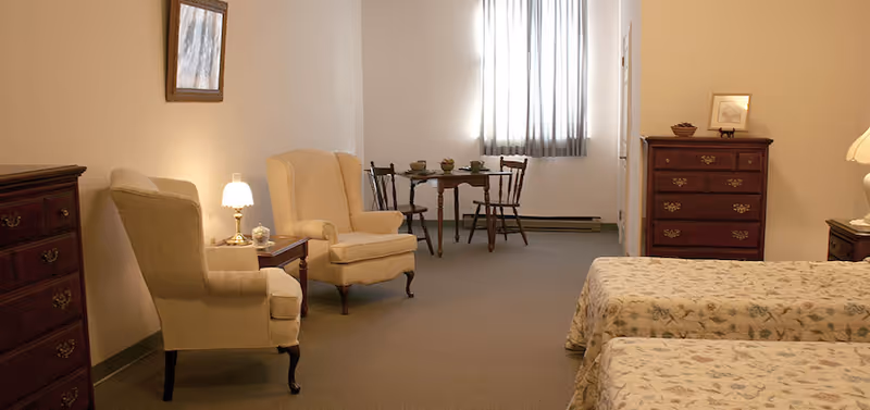 Senior living room with two beds, armchairs, and a small dining table in a bright bedroom.