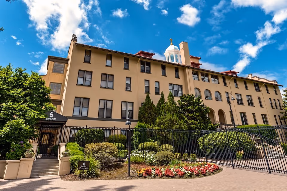 Exterior view of a large beige multi-story building with many windows, surrounded by well-maintained greenery and colorful flower beds under a bright blue sky with scattered clouds. A black metal fence and gate are in front, with a sign indicating the main entrance to St. Andrews Memory Care.
