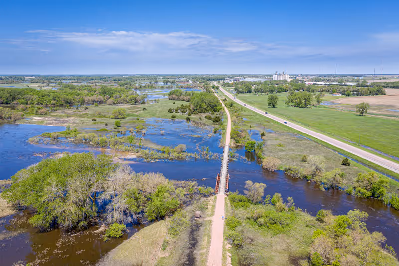 Aerial view of a river, trees, and a long bridge crossing open fields.