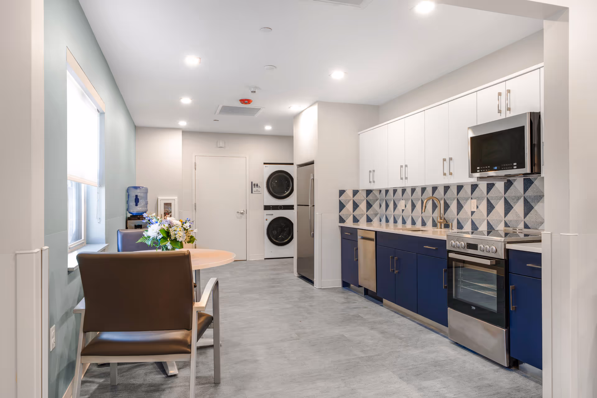 Modern facility kitchen with blue lower cabinets, white upper cabinets, a round table, and flowers.