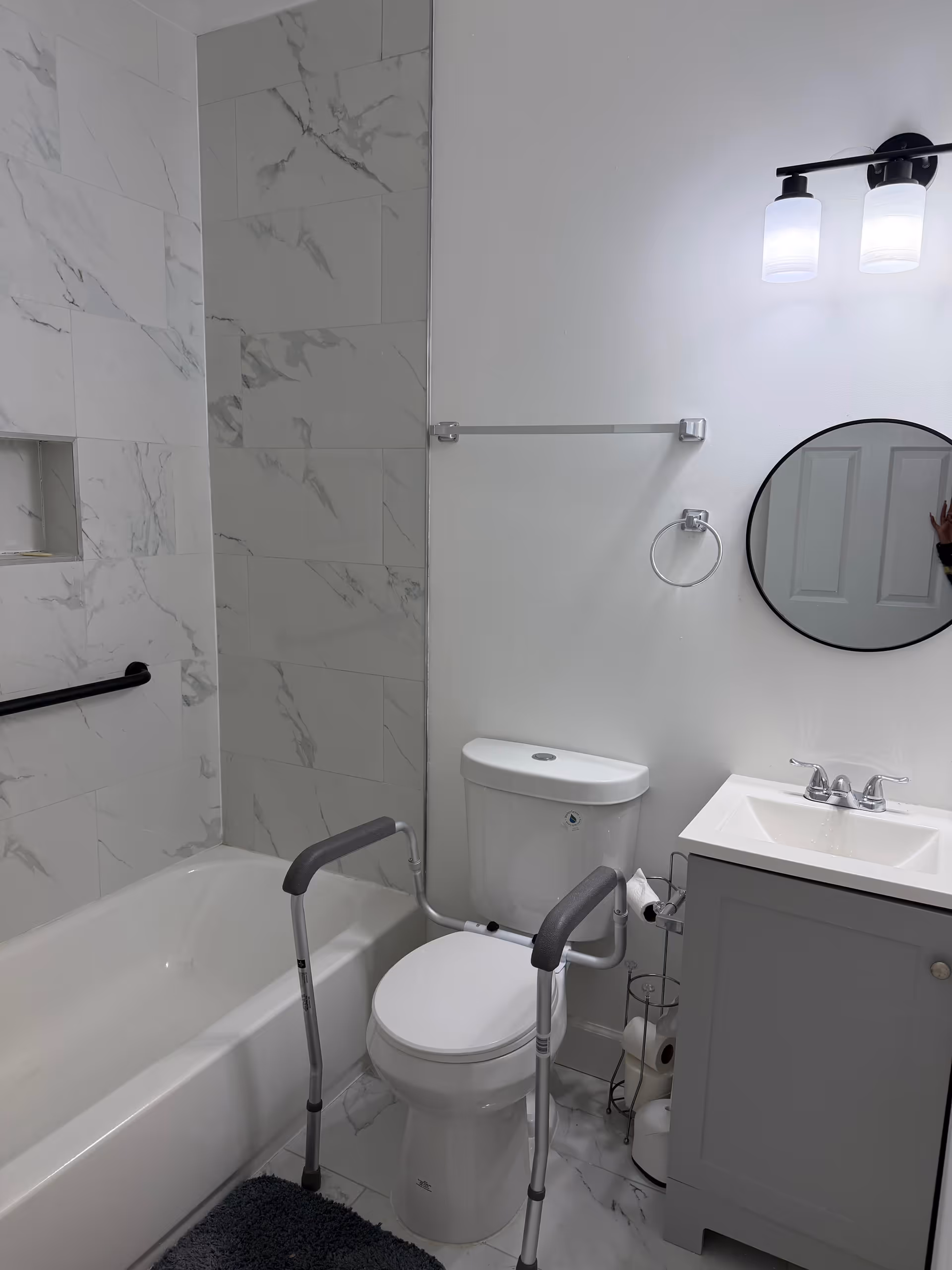 Accessible bathroom with tub, toilet, walker, and sink in marble-tiled space.