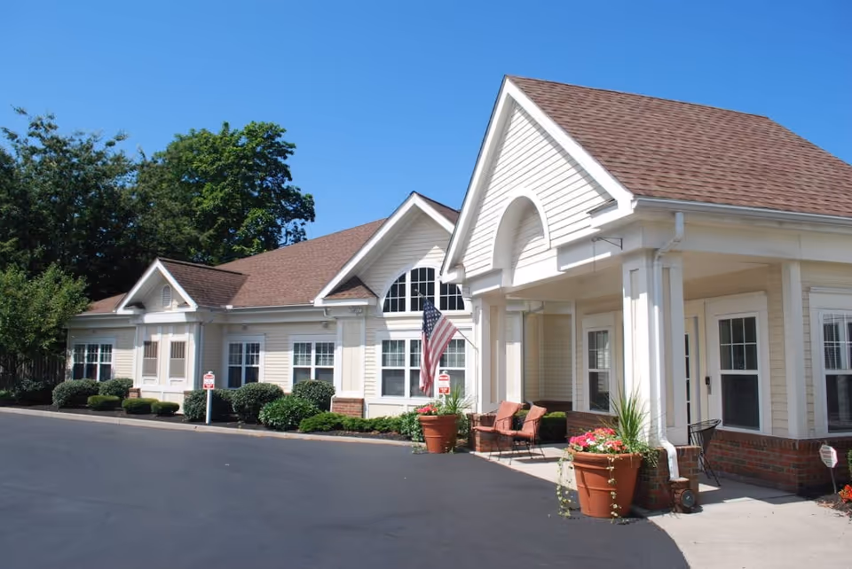 Front exterior of a single-story assisted living building with a covered entrance, American flag, potted plants and outdoor seating.