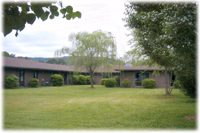 A grassy courtyard area with a few trees and bushes surrounded by a single-story brick building with a brown roof under a cloudy sky.
