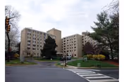 Wide exterior view of Sun Mar Nursing Center, a beige multi-story building with trees and crosswalk.