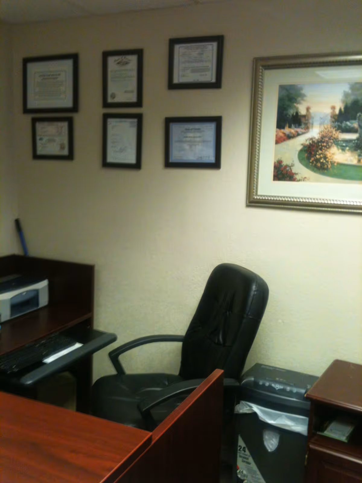 Office desk area with a black chair, printer, and framed certificates on the wall.