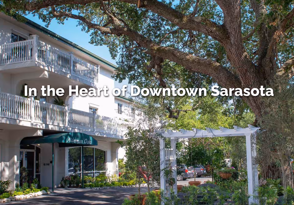 Exterior front of a white multi-story retirement residence with balconies, a green awning reading "Alderman Oaks", a large oak tree, and a pergola with landscaping.