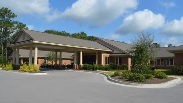 Front exterior of a single-story brick assisted living building with a covered porte-cochere, driveway and landscaped grounds under a blue sky.