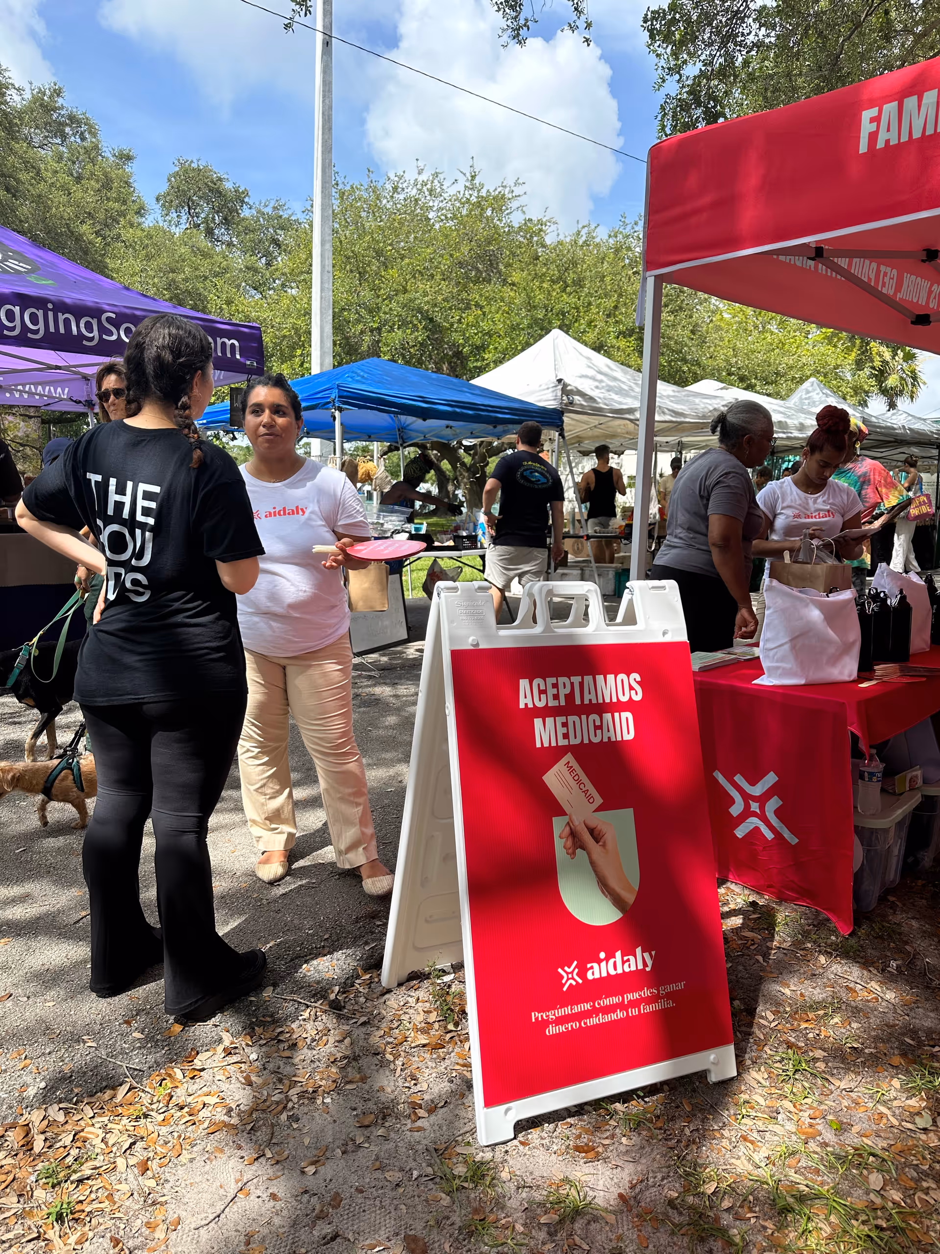 Outdoor community fair with tents and people; a red Aidaly Medicaid sign dominates foreground.