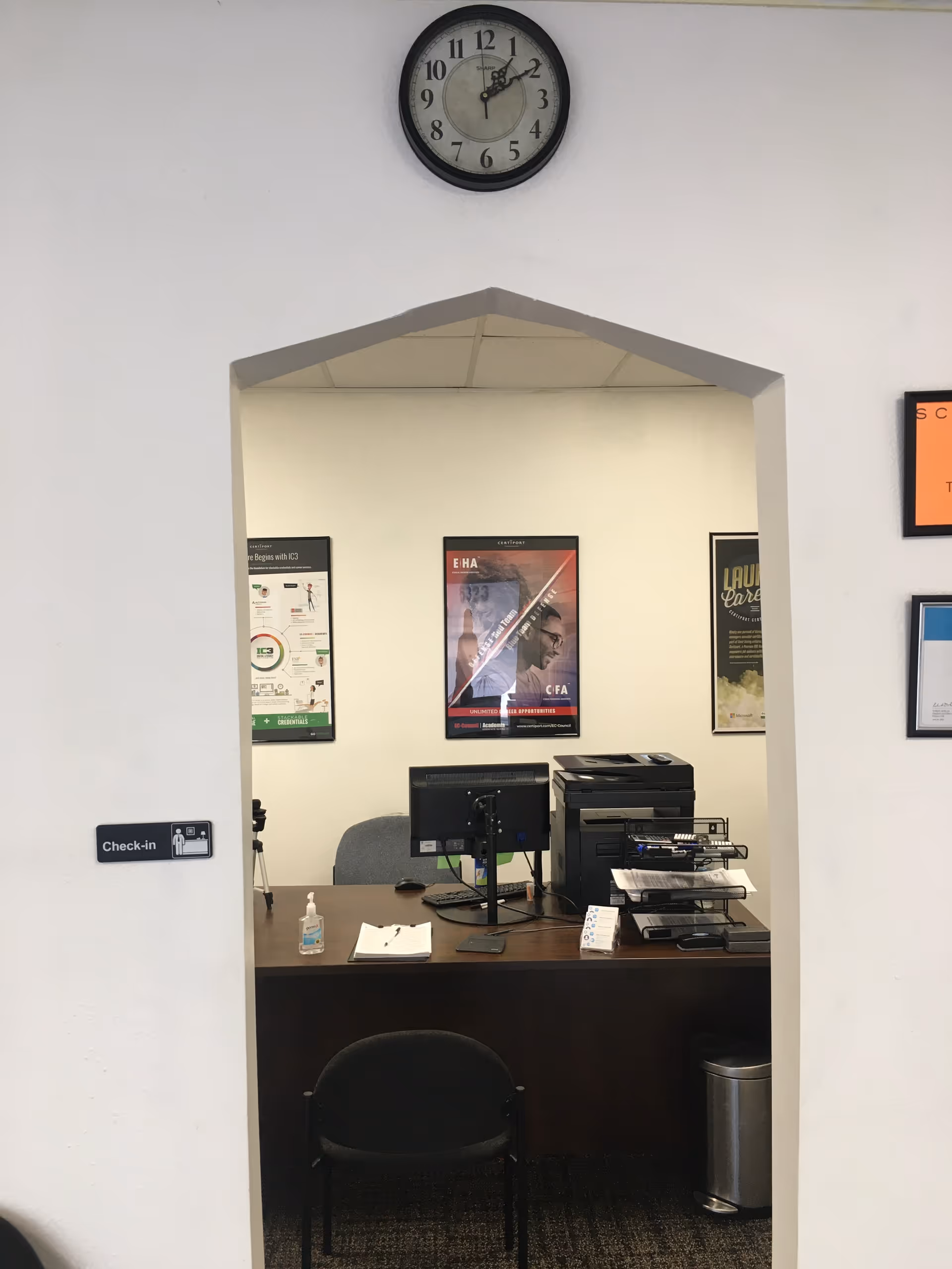 Reception desk in interior facility with check-in sign, computer, posters, and wall clock.