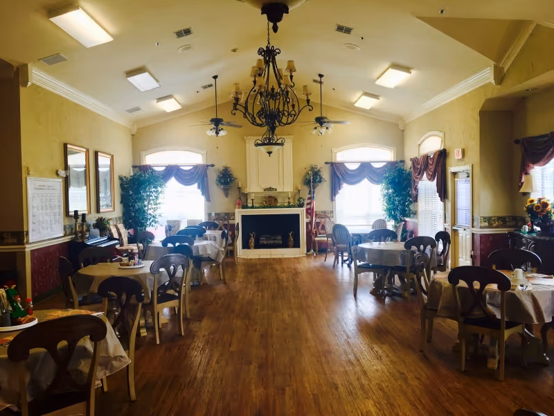 Spacious dining room with round tables and chairs, a central chandelier, fireplace, and large draped windows.