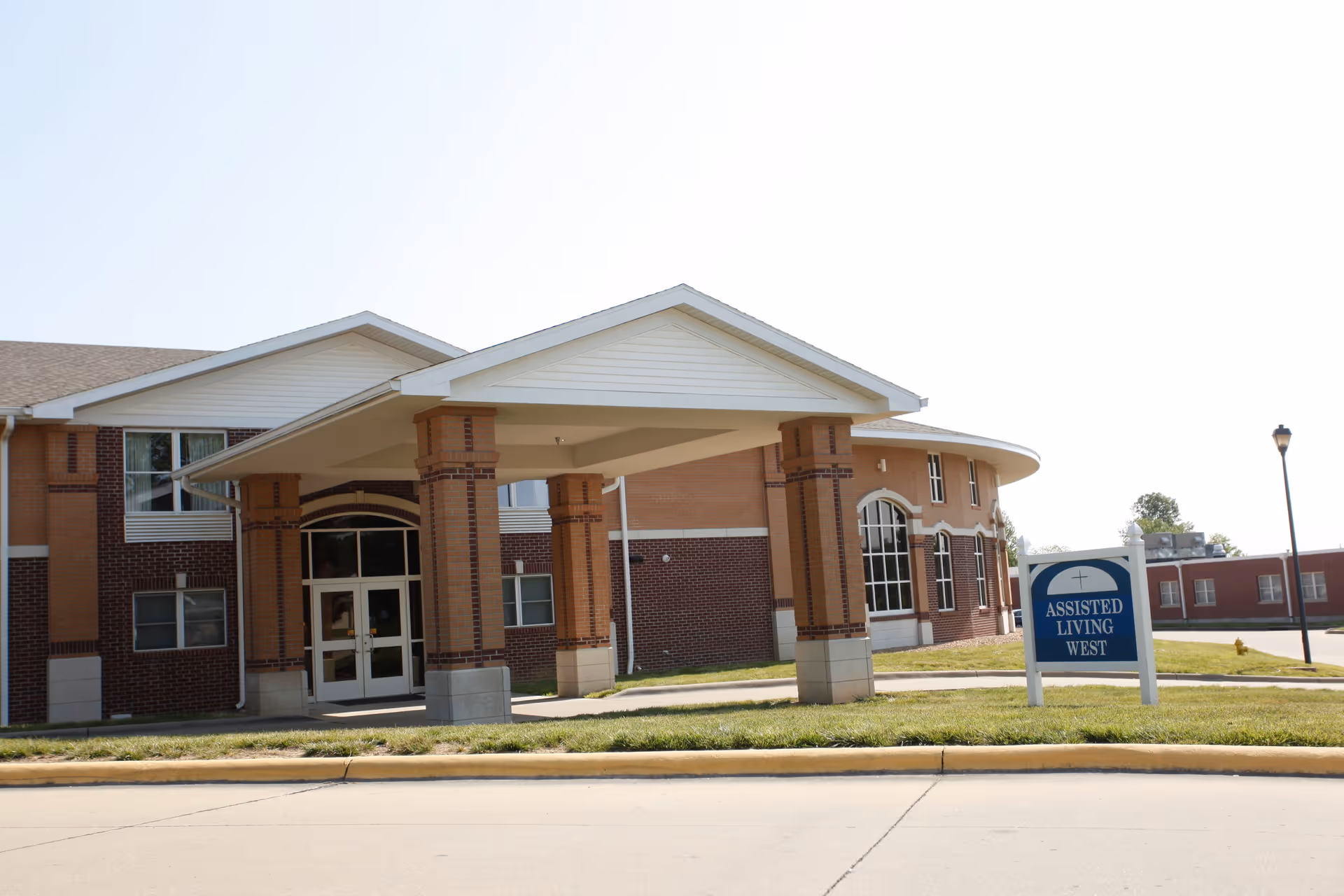 Front entrance of a brick assisted living building with a covered porte-cochère and a sign reading "Assisted Living West".