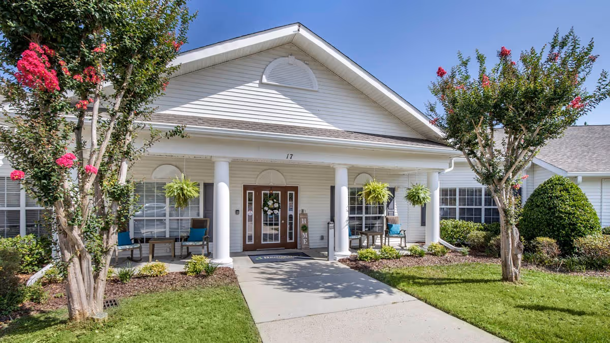 Front entrance of a single-story white senior living building with a covered porch supported by columns, hanging plants, seating, a concrete walkway, and landscaped trees and lawn.