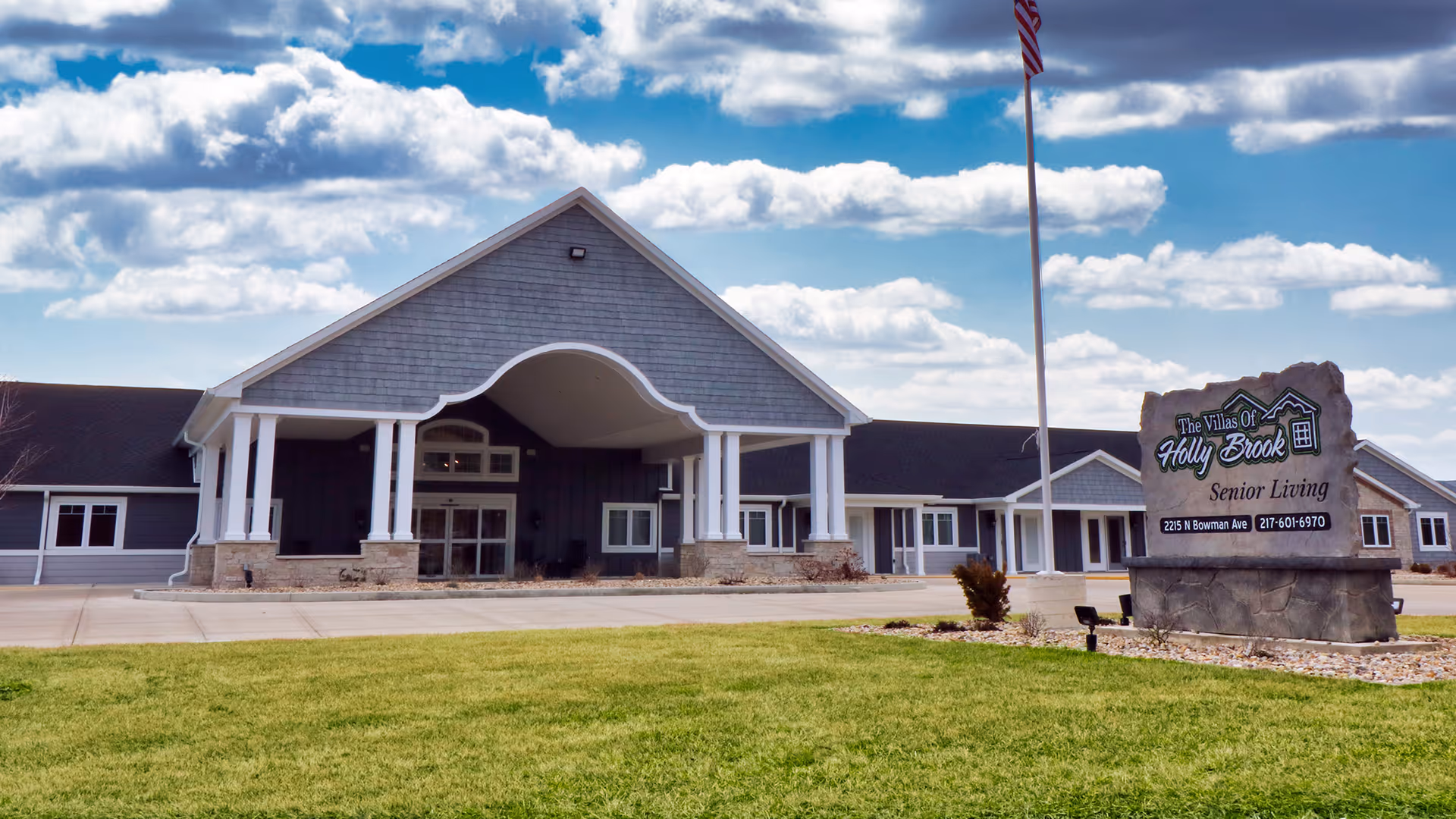 Exterior view of Villas of Holly Brook Assisted Living & Memory Care facility in Danville, IL, showing a large building with a covered entrance, white columns, and a stone sign with the facility name and contact information. The sky is partly cloudy and there is a well-maintained lawn in the foreground.
