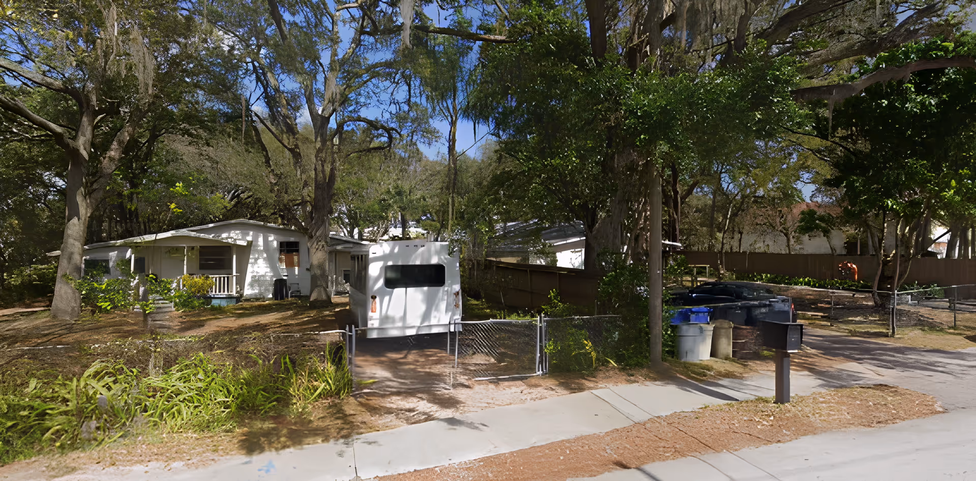 Single-story white house set among large trees with a driveway holding a white van and mailboxes and trash bins at the street.