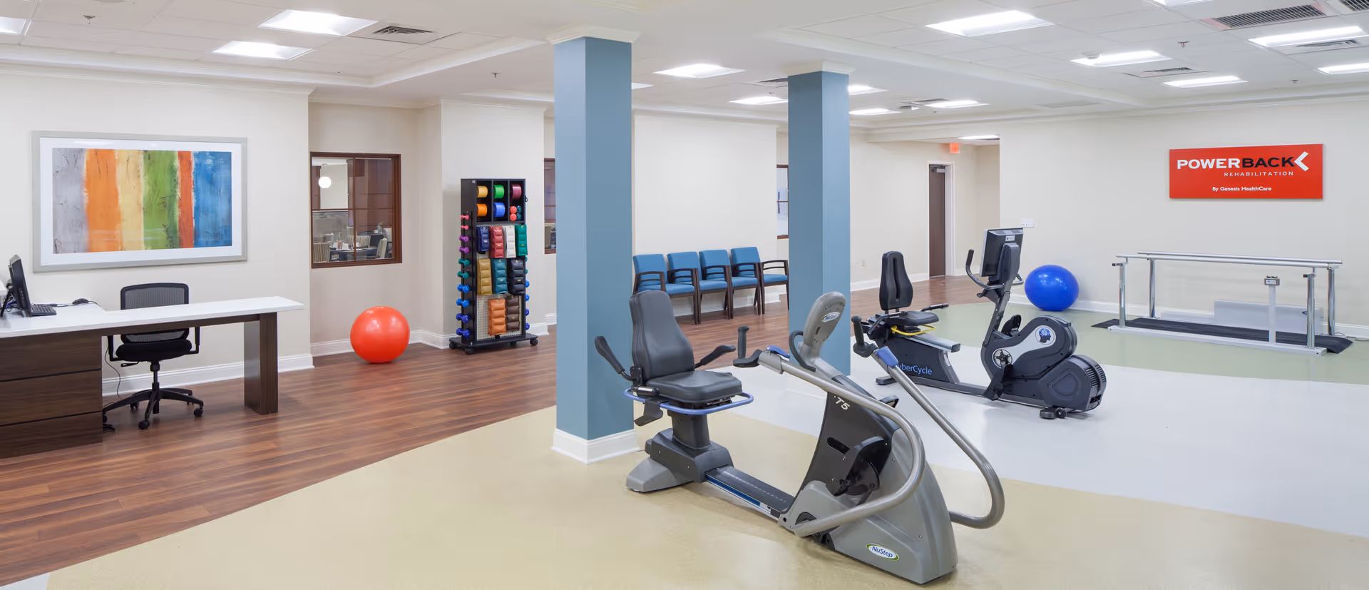 A rehabilitation exercise room with stationary exercise bikes, a treadmill, a rack of colorful dumbbells, exercise balls, a desk with a computer, and chairs along the wall. The room has light-colored walls, wood and vinyl flooring, and a red sign on the wall that reads 'POWERBACK Rehabilitation'.