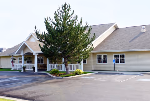 Front entrance of a single-story senior living building with a covered porch, a tree and a parking area with handicap spaces.