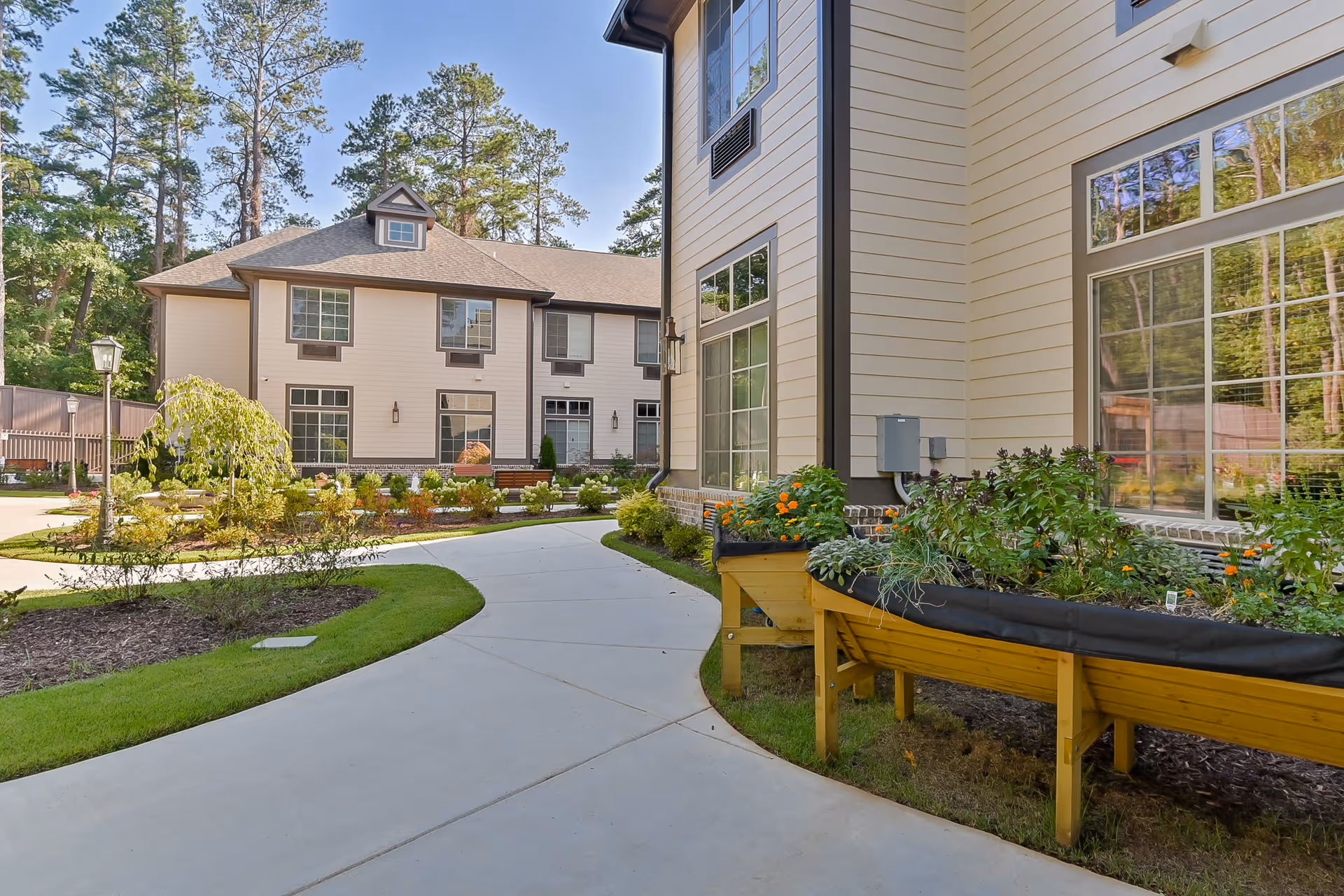 Curved paved walkway through a landscaped courtyard beside the exterior of a multi-story senior living building with raised garden beds.