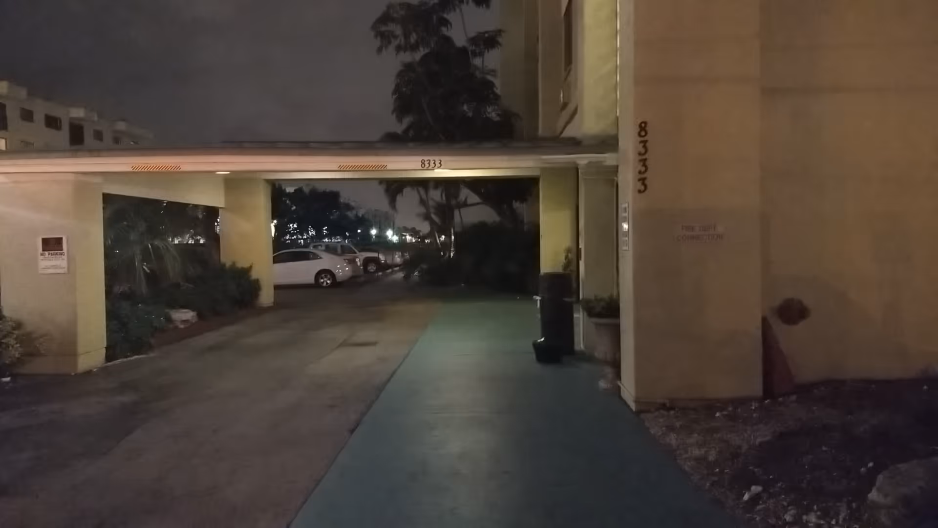 Night exterior view of a covered driveway and building entrance with parked cars.