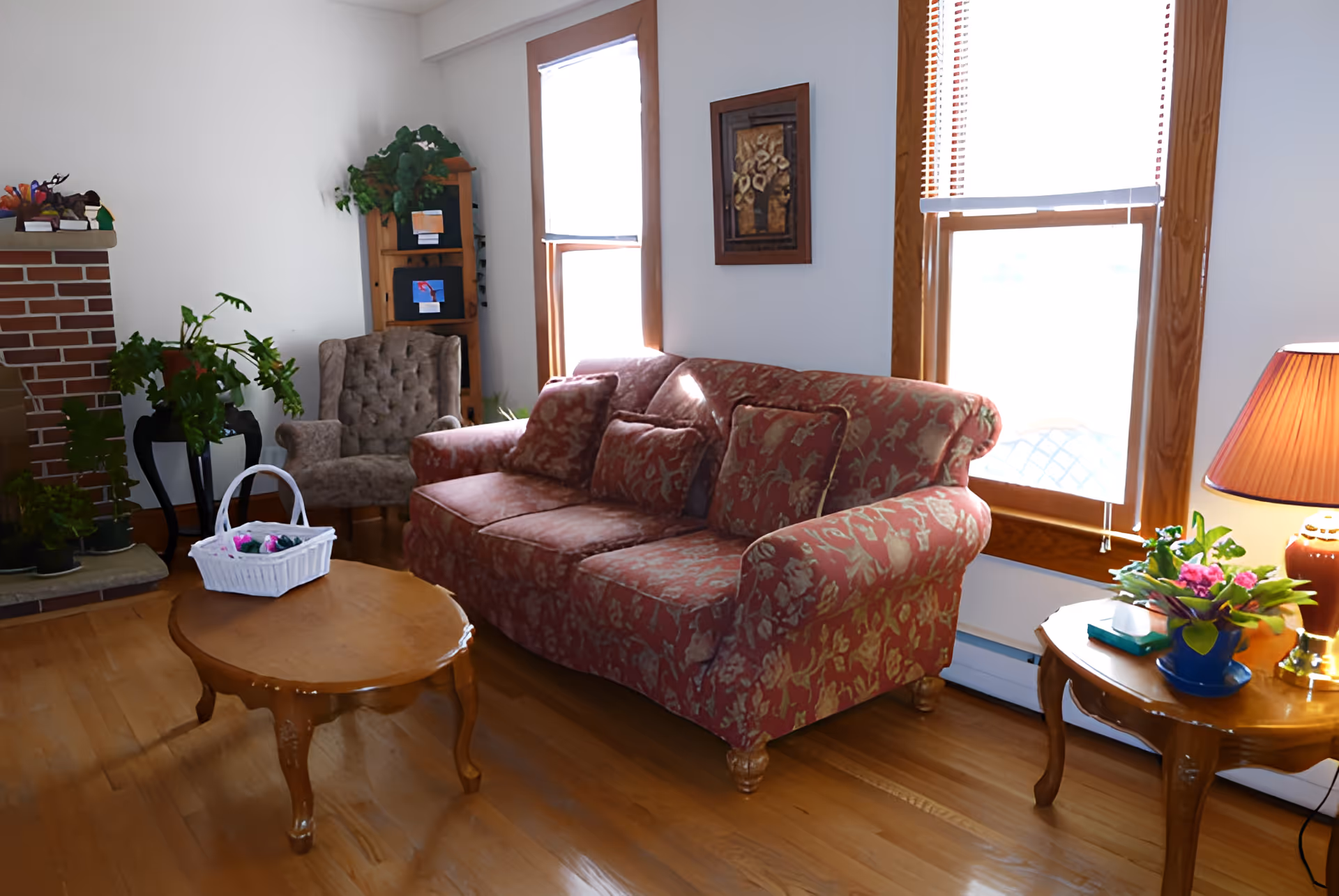 Sunlit living room with a patterned sofa, wooden coffee and side tables, armchair, plants, and two windows.