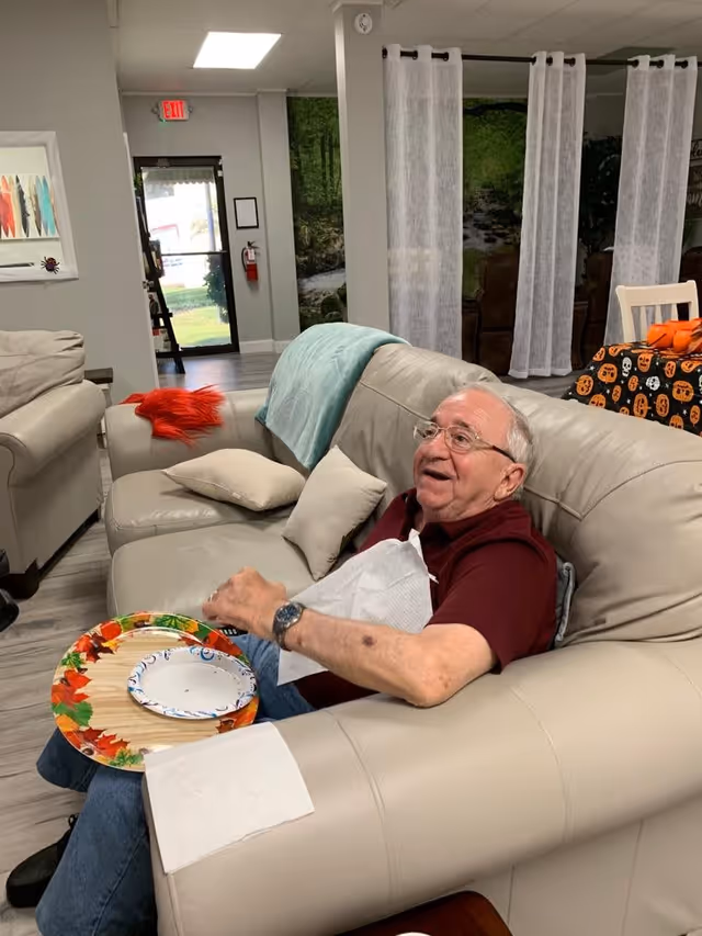 An elderly man wearing glasses and a maroon shirt is sitting on a beige leather couch in a cozy room. He has a paper napkin tucked into his shirt and is holding a large plate with a smaller plate on top, both decorated with autumn leaves. Behind him, there is another beige couch with a red throw and a light blue blanket draped over it. In the background, there are white sheer curtains, a table covered with a Halloween-themed tablecloth, and a door leading outside.
