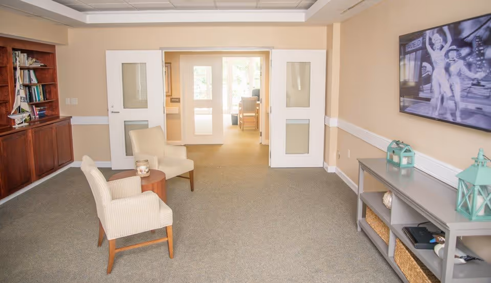 A bright and spacious living room area in an assisted living facility featuring two beige armchairs around a small wooden table, a built-in wooden bookshelf filled with books and decorative items on the left, and a gray console table with decorative lanterns and baskets on the right. A large flat-screen TV is mounted on the wall above the console table, displaying a black and white image. Double doors lead to another room with natural light coming through windows.