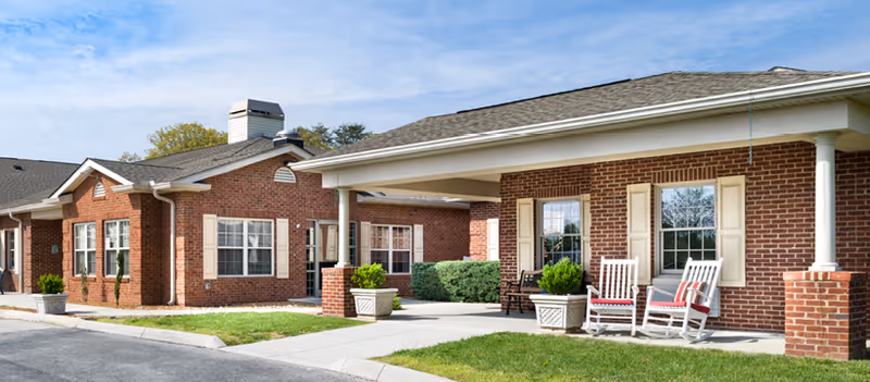 Brick assisted living facility exterior with a covered entrance and rocking chairs on a patio.