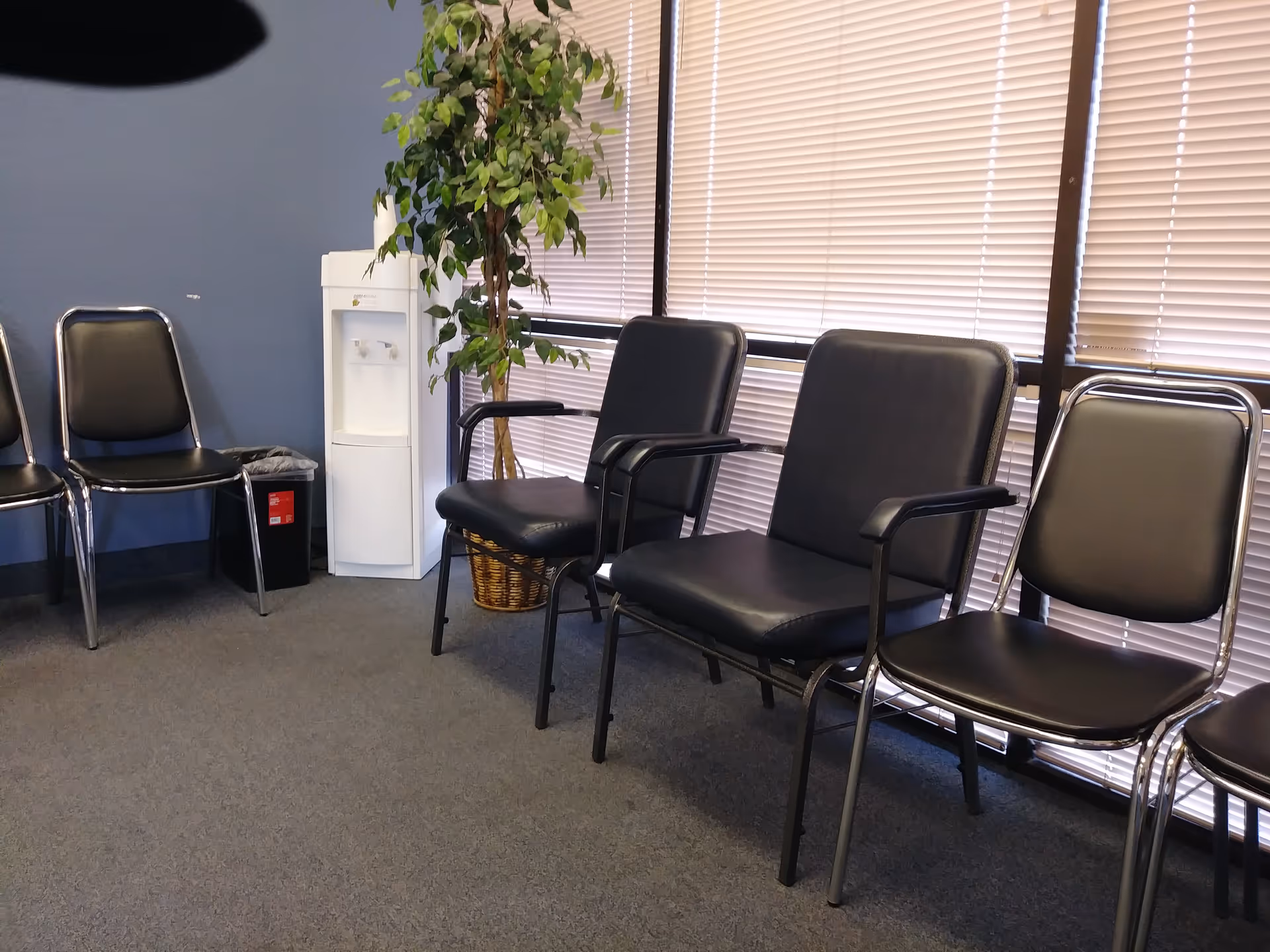 A waiting area with black chairs, blinds, a water cooler, and a plant