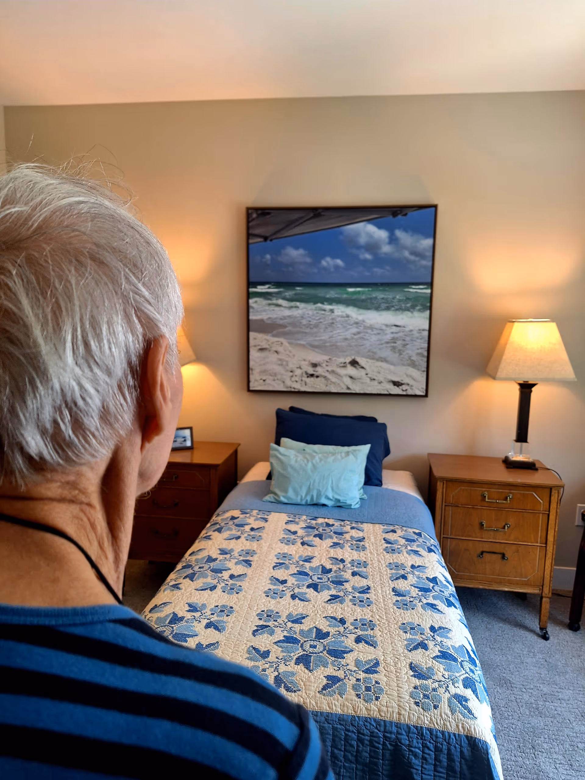 Bedroom in a senior living facility with bed, nightstands, lamps, and wall art.