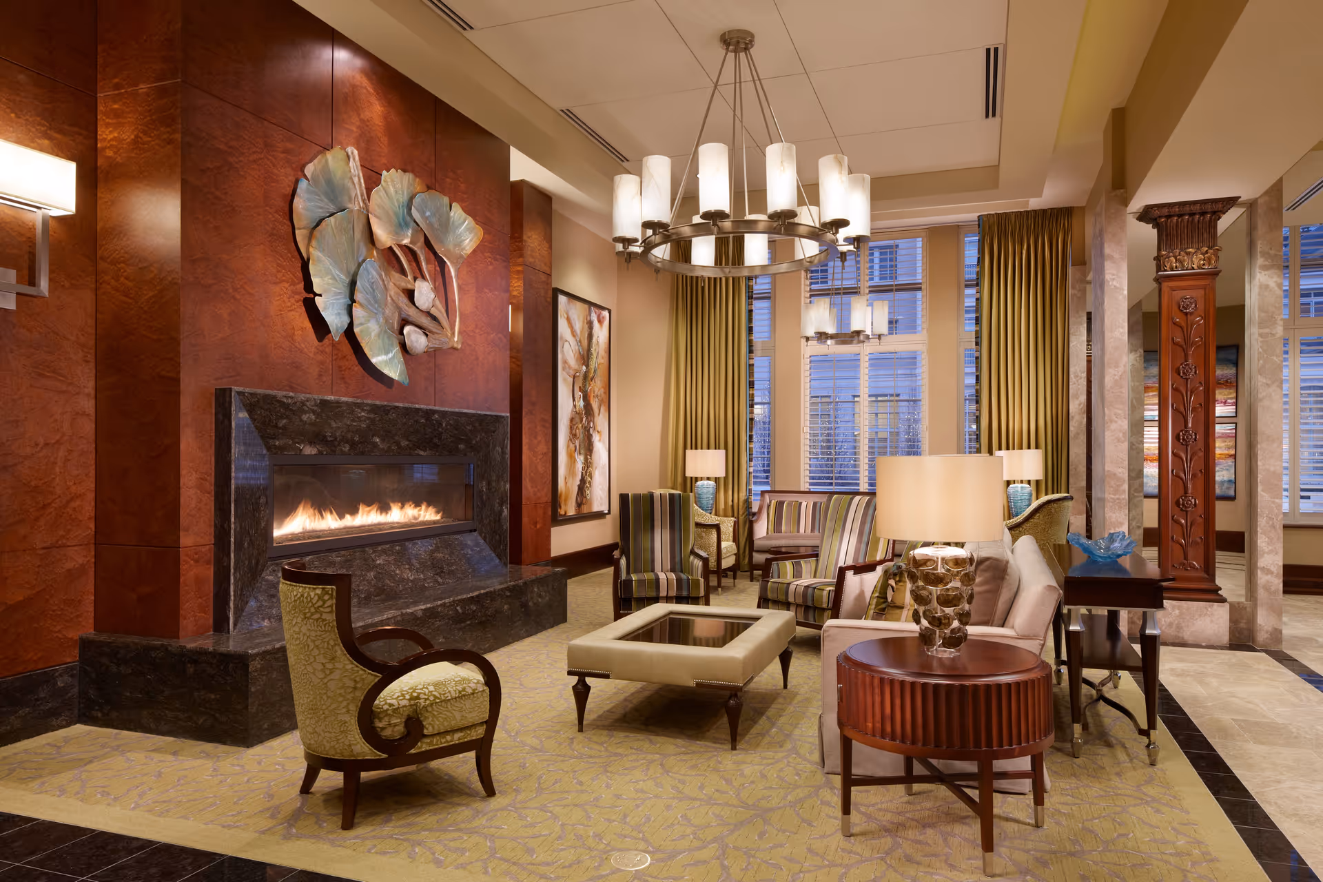 A cozy and elegant living room area with a modern fireplace set in a dark marble surround against a rich wooden wall. The room features a variety of seating options including upholstered armchairs and a sofa, arranged around a square coffee table. Large windows with gold curtains allow natural light to fill the space. Decorative elements include a large metal wall sculpture above the fireplace, a chandelier with cylindrical lampshades, and a wooden column with intricate carvings.