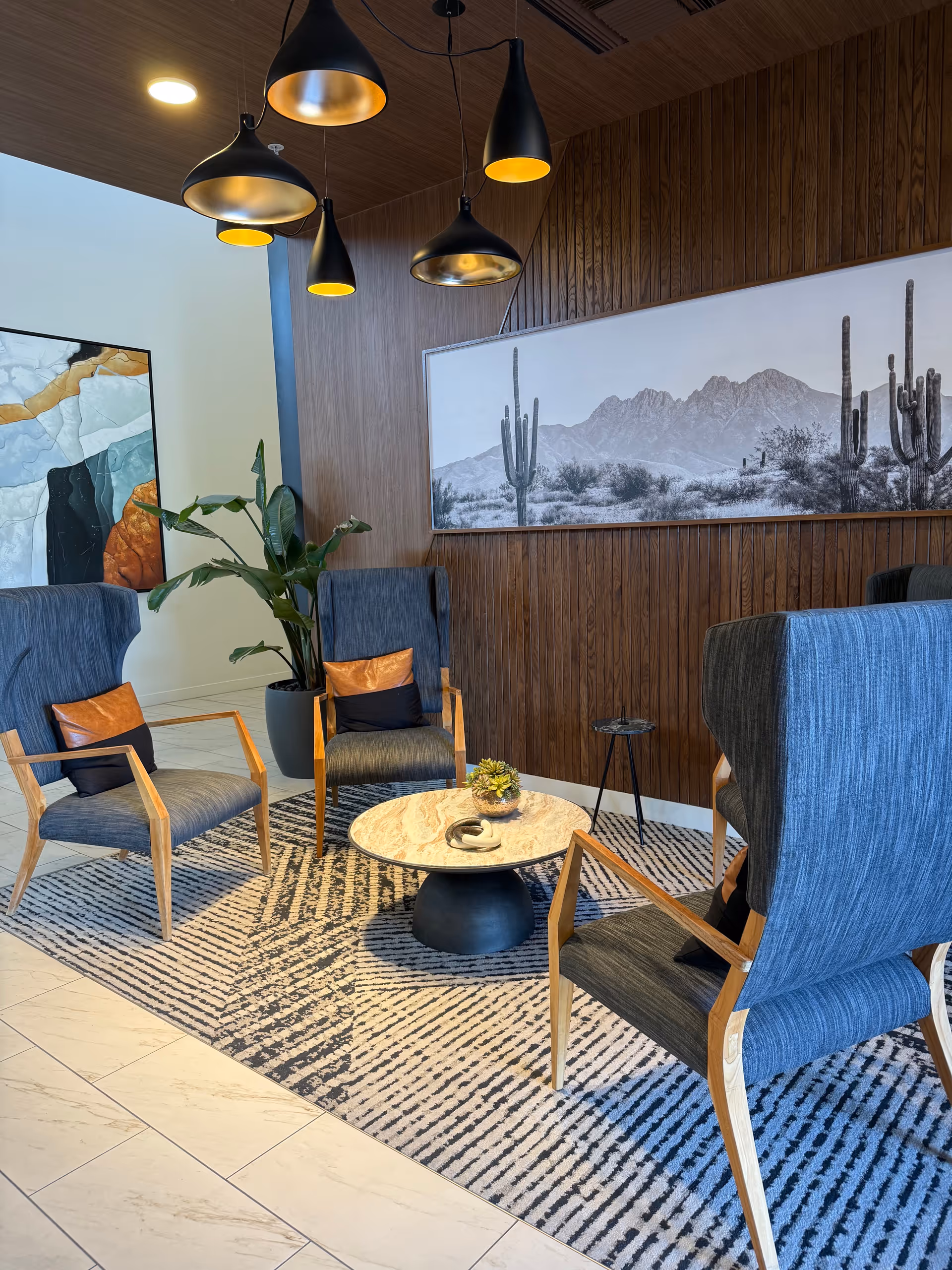 Modern lounge with blue chairs, wooden wall, desert artwork, and pendant lighting.
