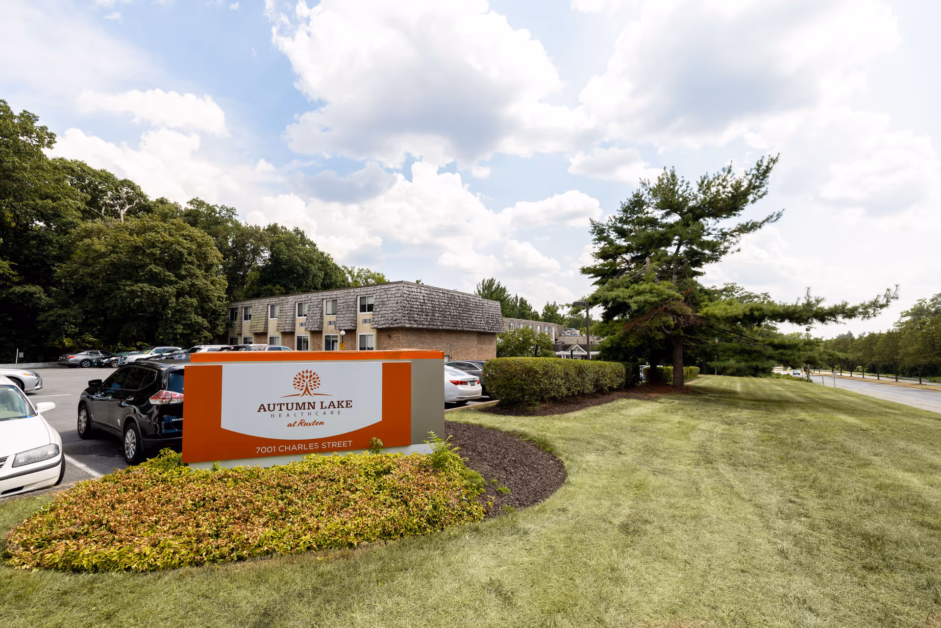 Exterior view of Autumn Lake Healthcare at Ruxton facility showing a parking lot with several cars, a large sign with the facility name and address, a grassy area with trees, and a partly cloudy sky.