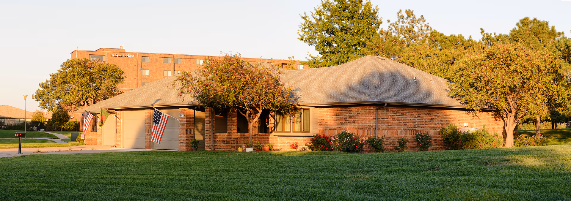 A single-story brick building with a gray shingled roof surrounded by green grass and trees, with American flags displayed near the entrance. In the background, a taller brick building with the sign 'Presbyterian Manor' is visible.