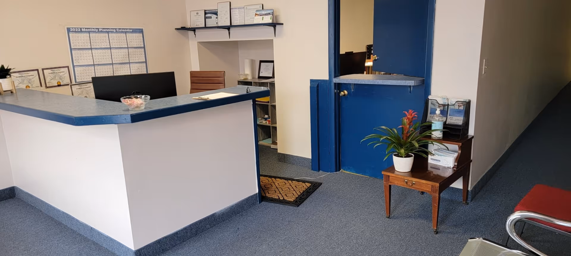 Reception desk in a senior living facility's interior with blue trim, shelving, and a potted plant.