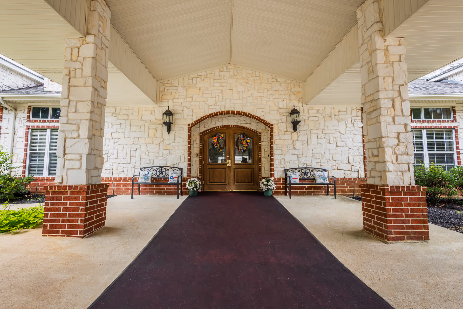 Covered entrance to a senior living facility with stone and brick pillars, a maroon carpet leading to double wooden doors decorated with wreaths, two black benches with pillows on either side, and potted plants near the doors.
