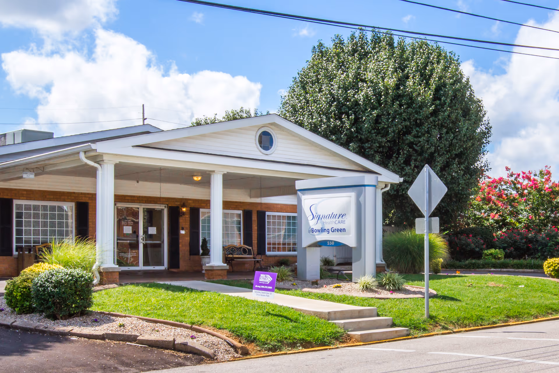 Exterior front view of Signature HealthCARE of Bowling Green building with a covered entrance supported by white columns, a sign displaying the facility name, green lawn, bushes, and a large tree under a partly cloudy sky.