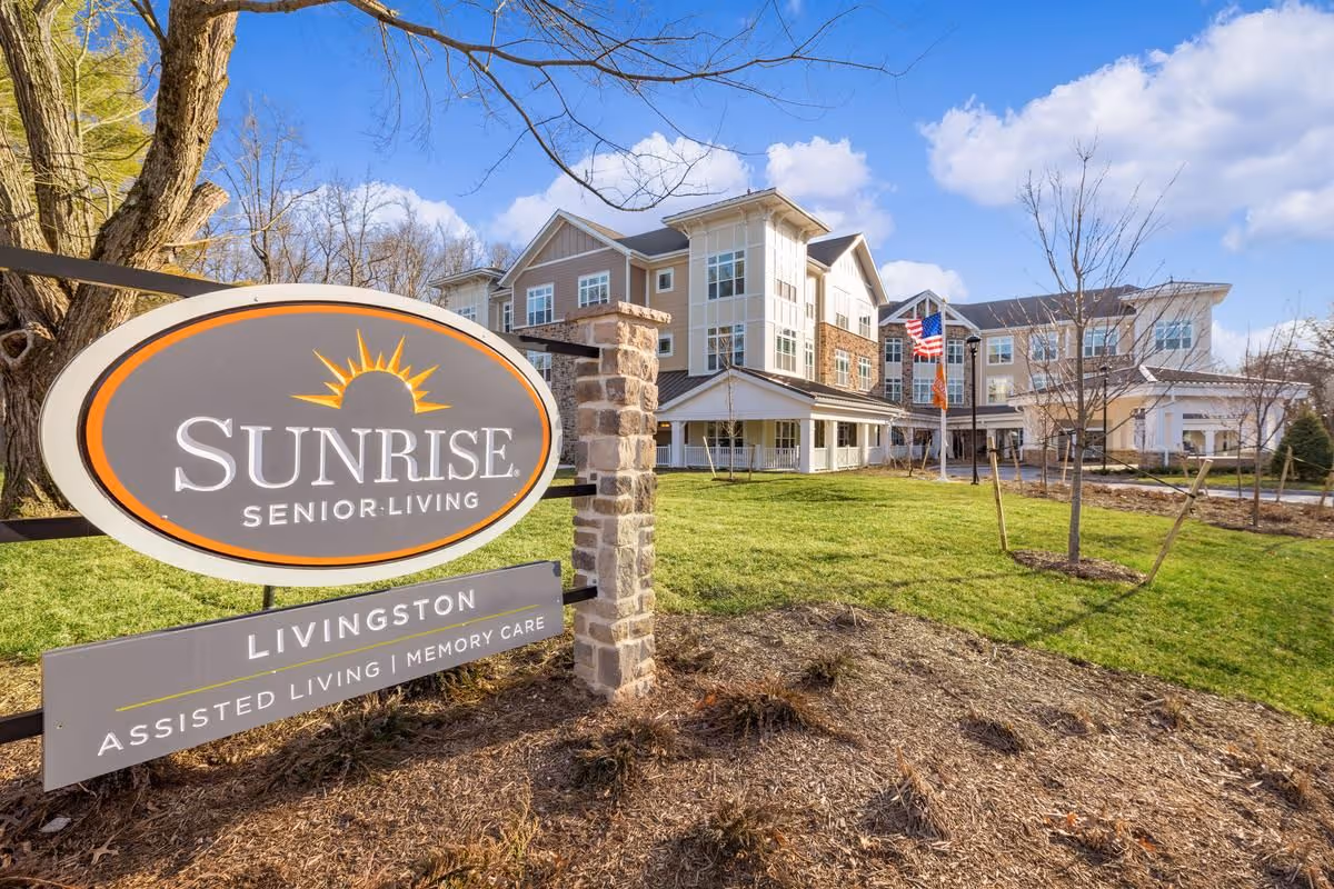 Exterior view of Sunrise Senior Living facility in Livingston, showing a large multi-story building with a porch, green lawn, trees, and an American flag. In the foreground, there is a sign with the Sunrise Senior Living logo and text indicating Assisted Living and Memory Care services.