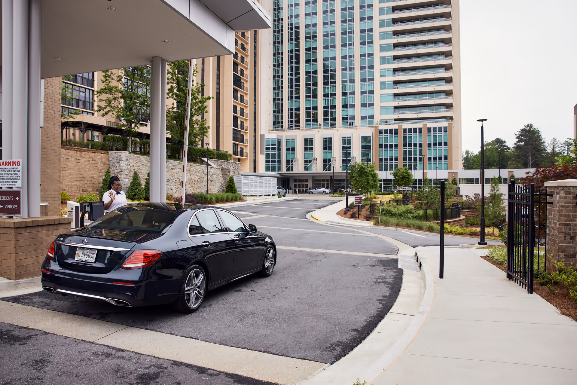 A black sedan parked at the drive-up entrance of a tall residential building with a security attendant at the gate.