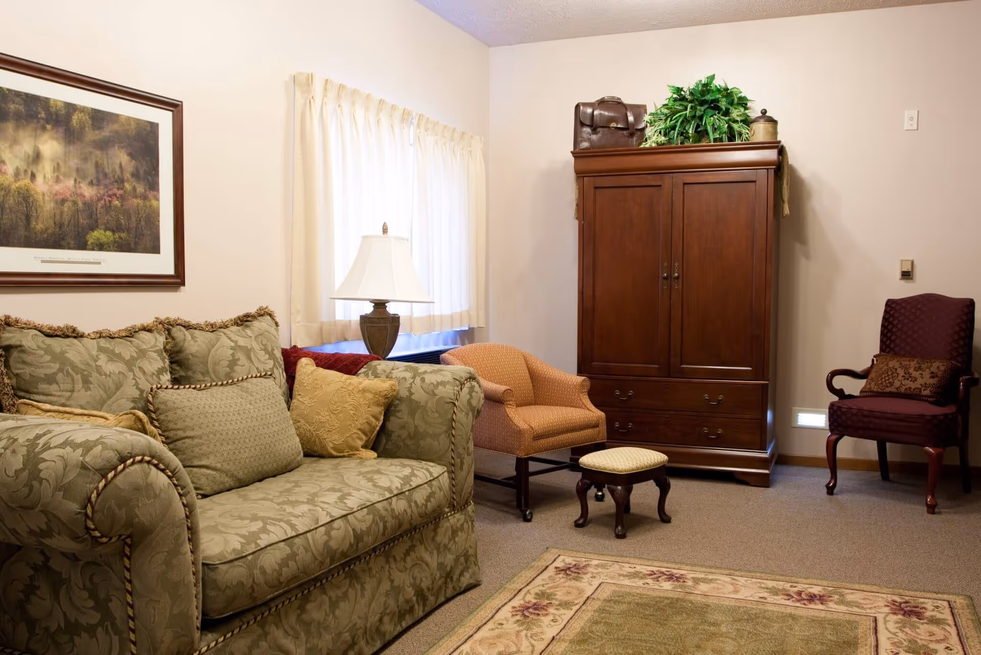 A cozy living room with a patterned green sofa adorned with multiple cushions, a small orange armchair with a matching footstool, a wooden armoire topped with a green plant and a brown bag, a burgundy armchair with a decorative pillow, a table lamp on a side table, a framed landscape painting on the wall, and a window with cream-colored curtains.