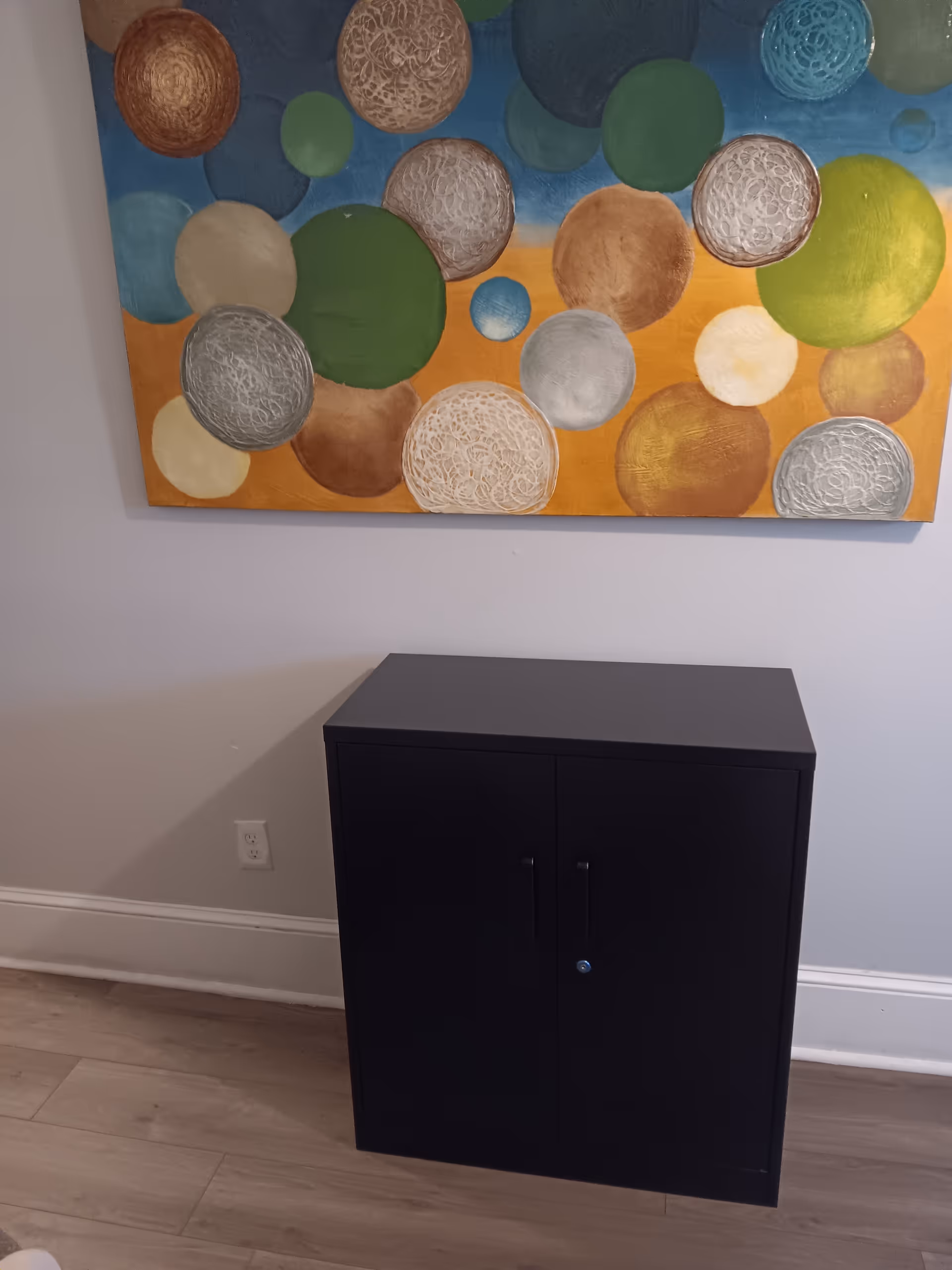 A black cabinet stands under a colorful abstract painting in an interior hallway.