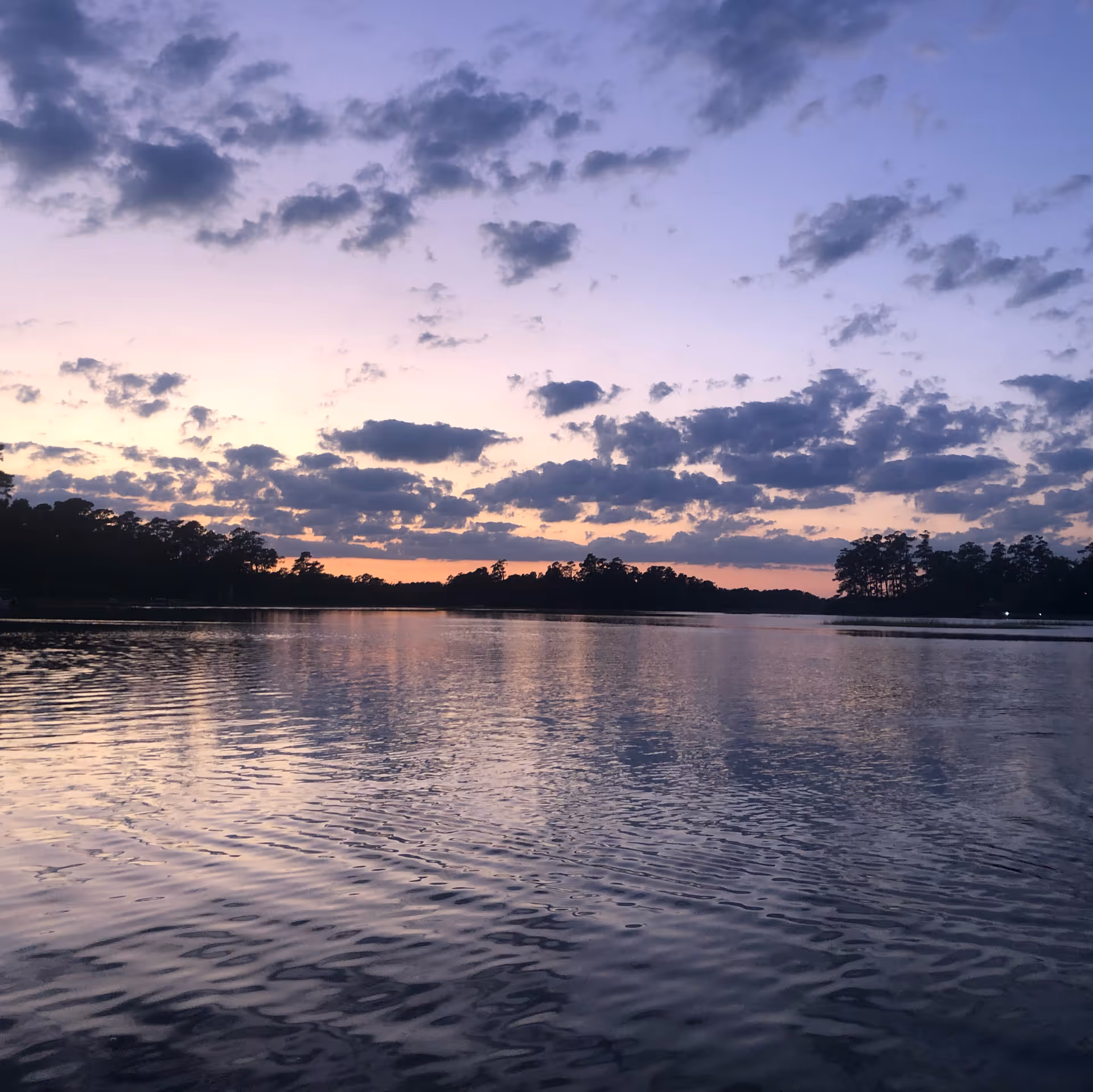 Calm lake at sunset with tree-lined shore and purple, cloudy sky