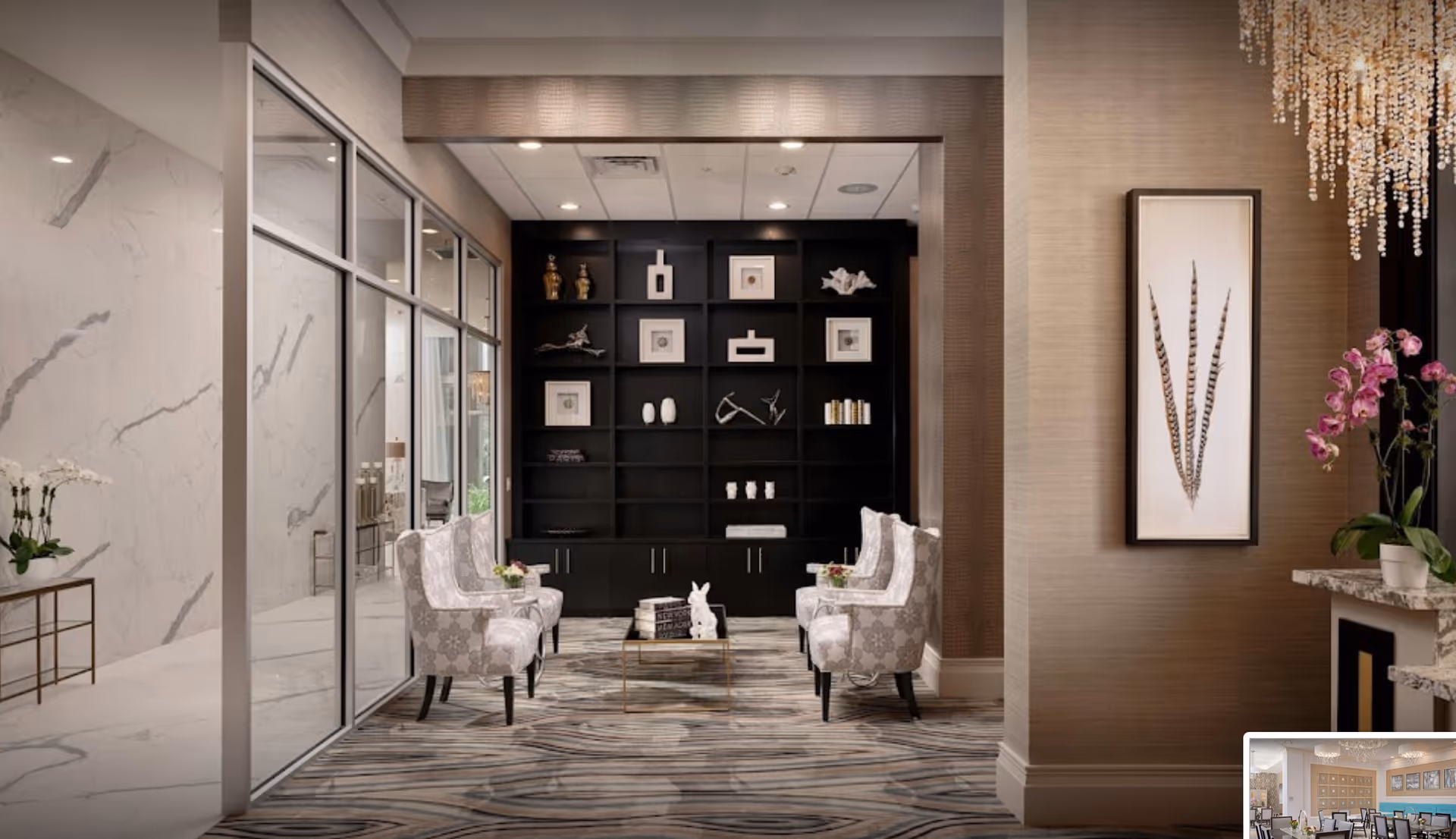 Elegant lounge area with patterned armchairs facing a black built-in bookshelf, marble walls, artwork and a chandelier.